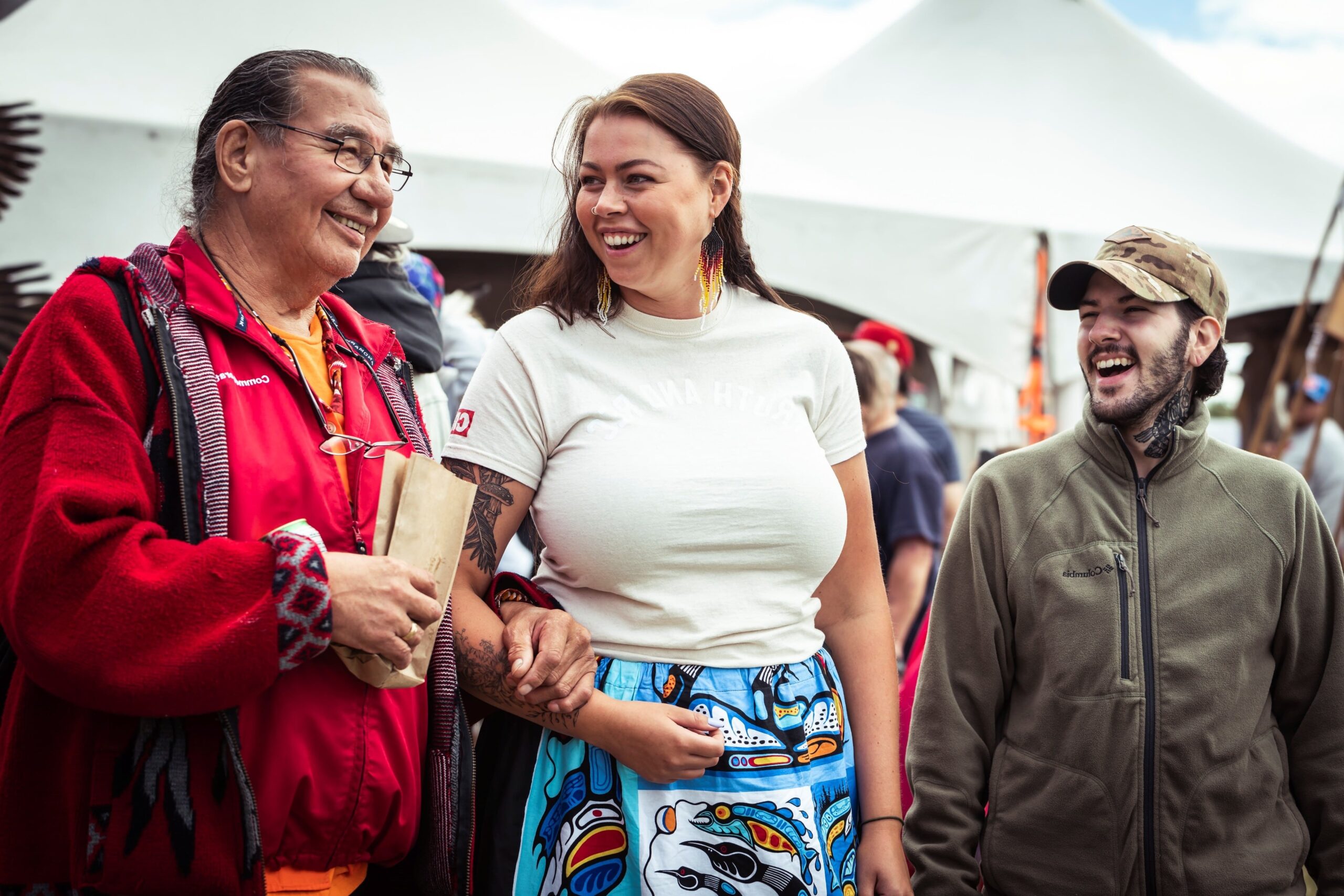 Un Aîné autochtone et deux jeunes lors d’un pow-wow en plein air, dans une ambiance festive et communautaire.