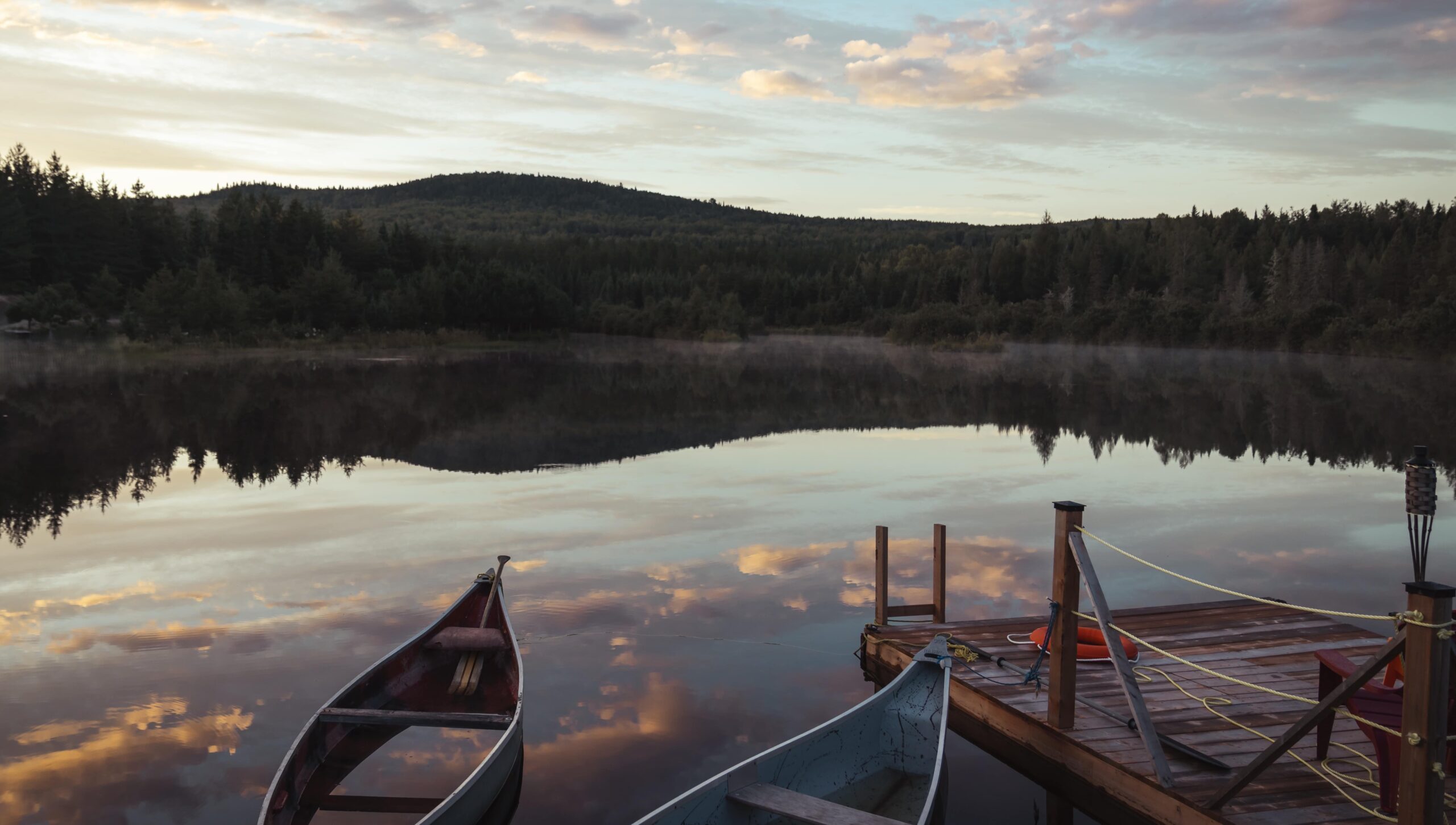 Deux canots amarrés à un quai en bois sur un lac calme au lever ou coucher du soleil, entouré de collines boisées.