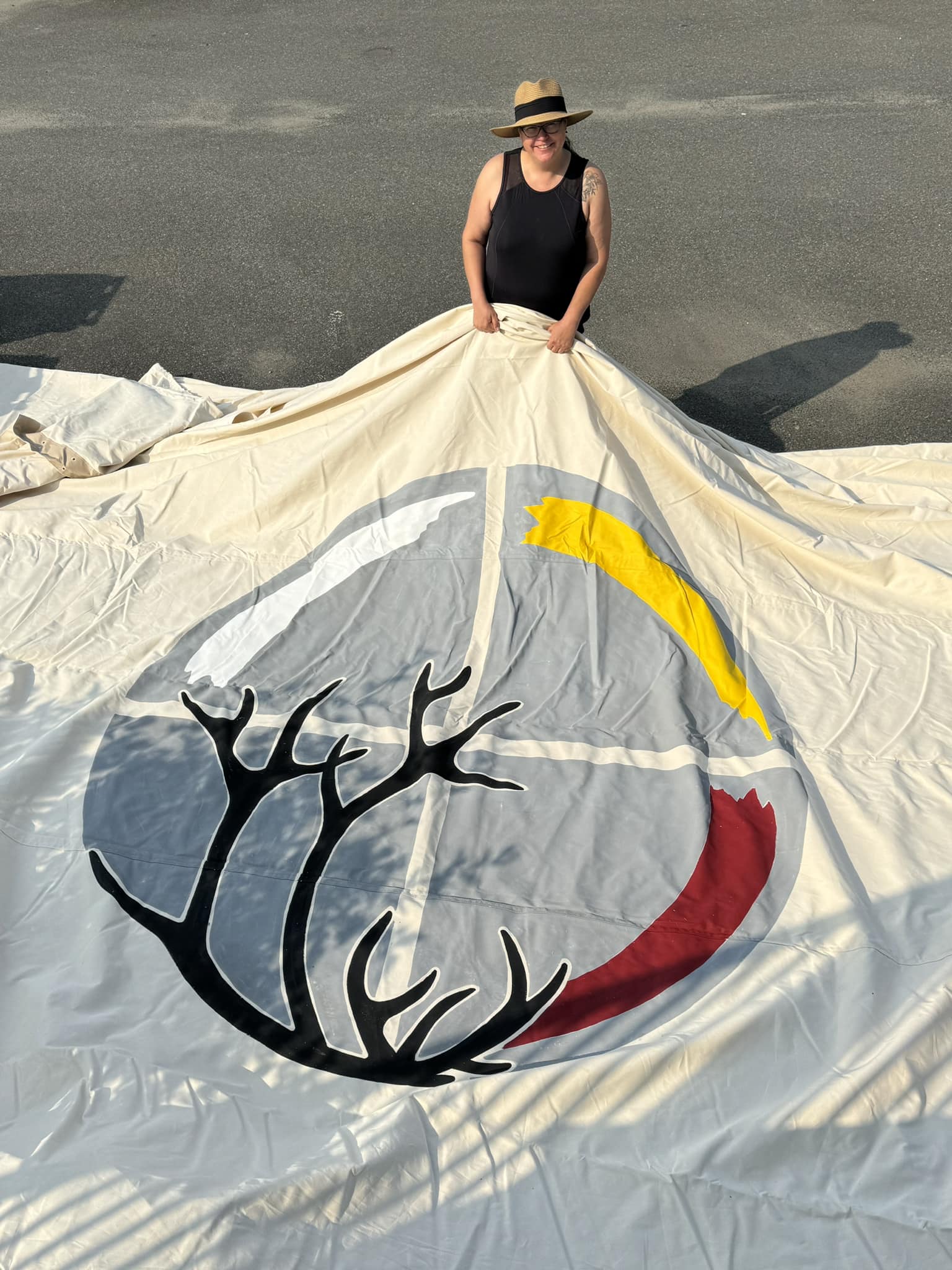 Grande toile avec le logo du Centre d'amitié autochtone de Sept-Îles (roue de médecine autochtone et des bois de cervidé) tenue à l’extérieur sur un sol asphalté.