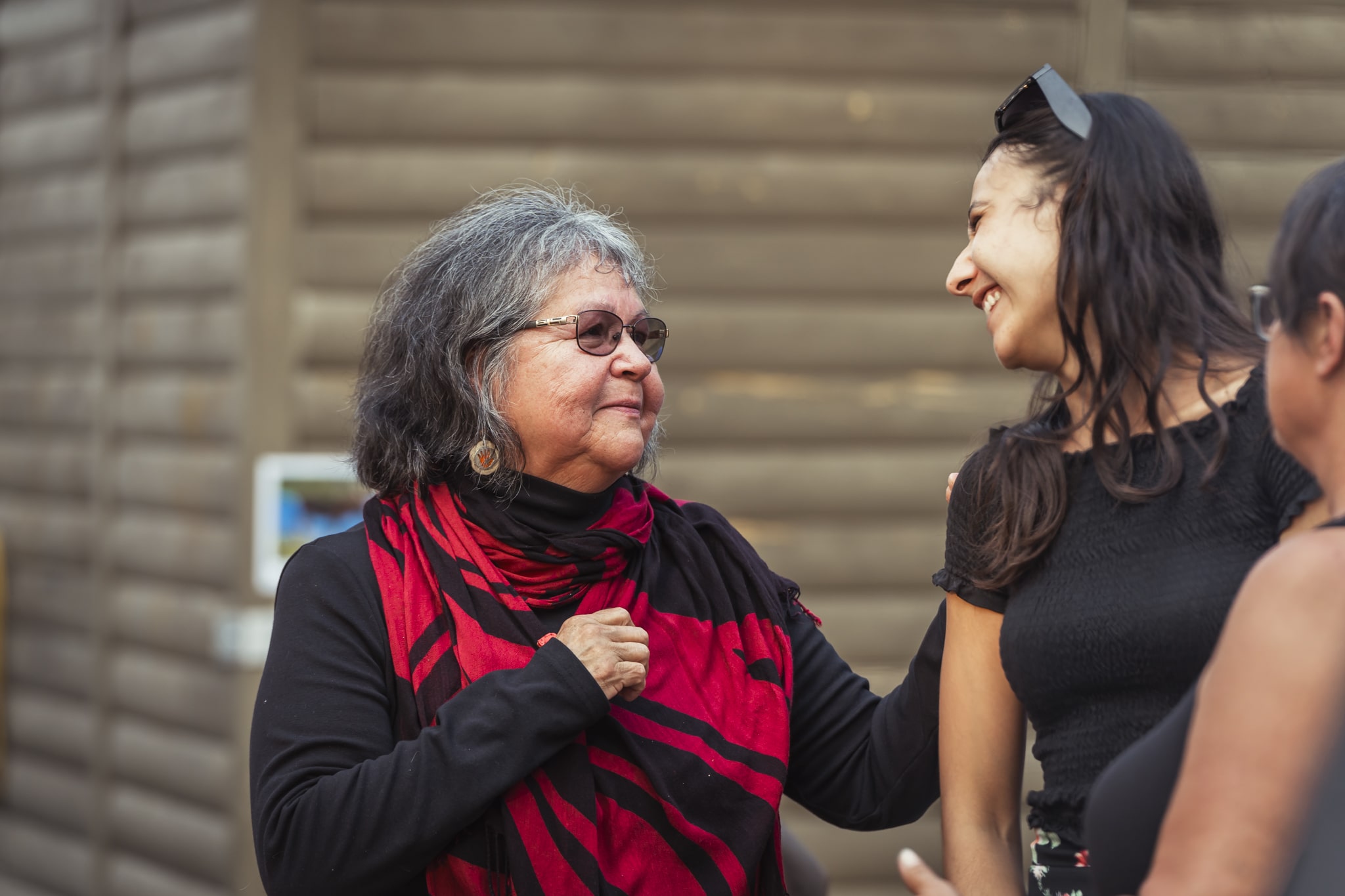 Aînée autochtone en interaction avec une jeune femme devant un bâtiment en bois.