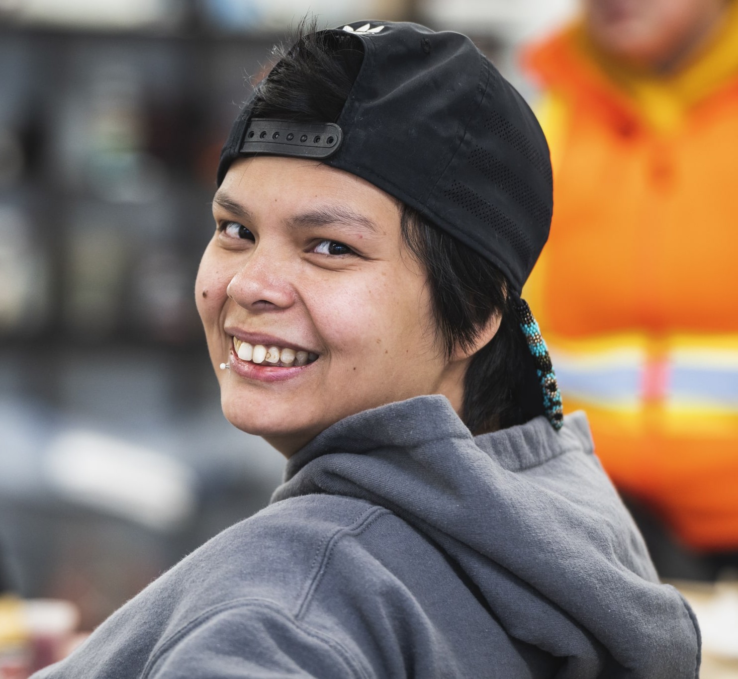 Personne autochtone souriante dans un centre d’amitié, vêtue d’un chandail à capuchon et d’une casquette.