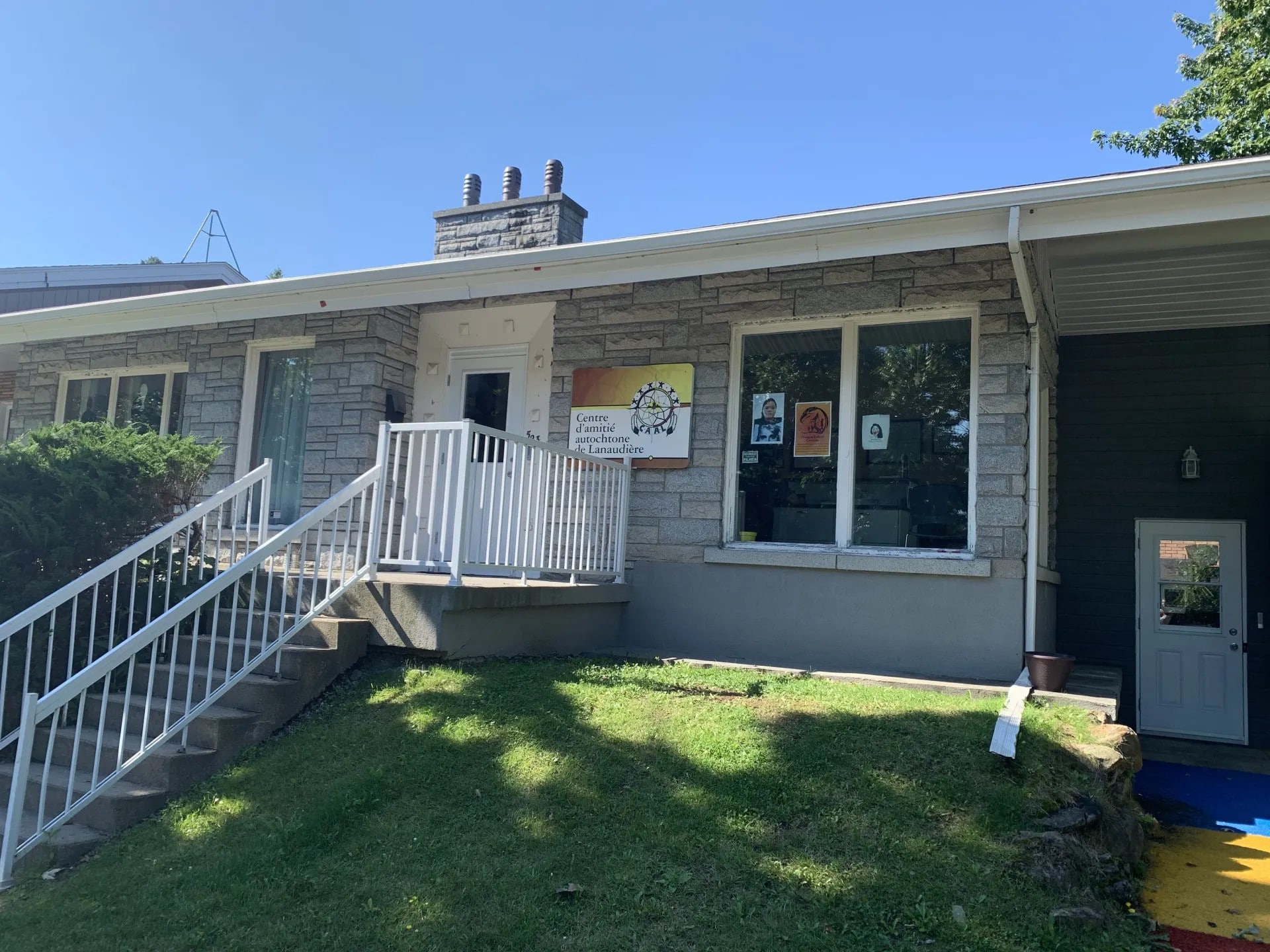 Façade en pierre du Centre d’amitié autochtone de Lanaudière avec entrée surélevée, rampes blanches et affiches visibles dans les fenêtres.