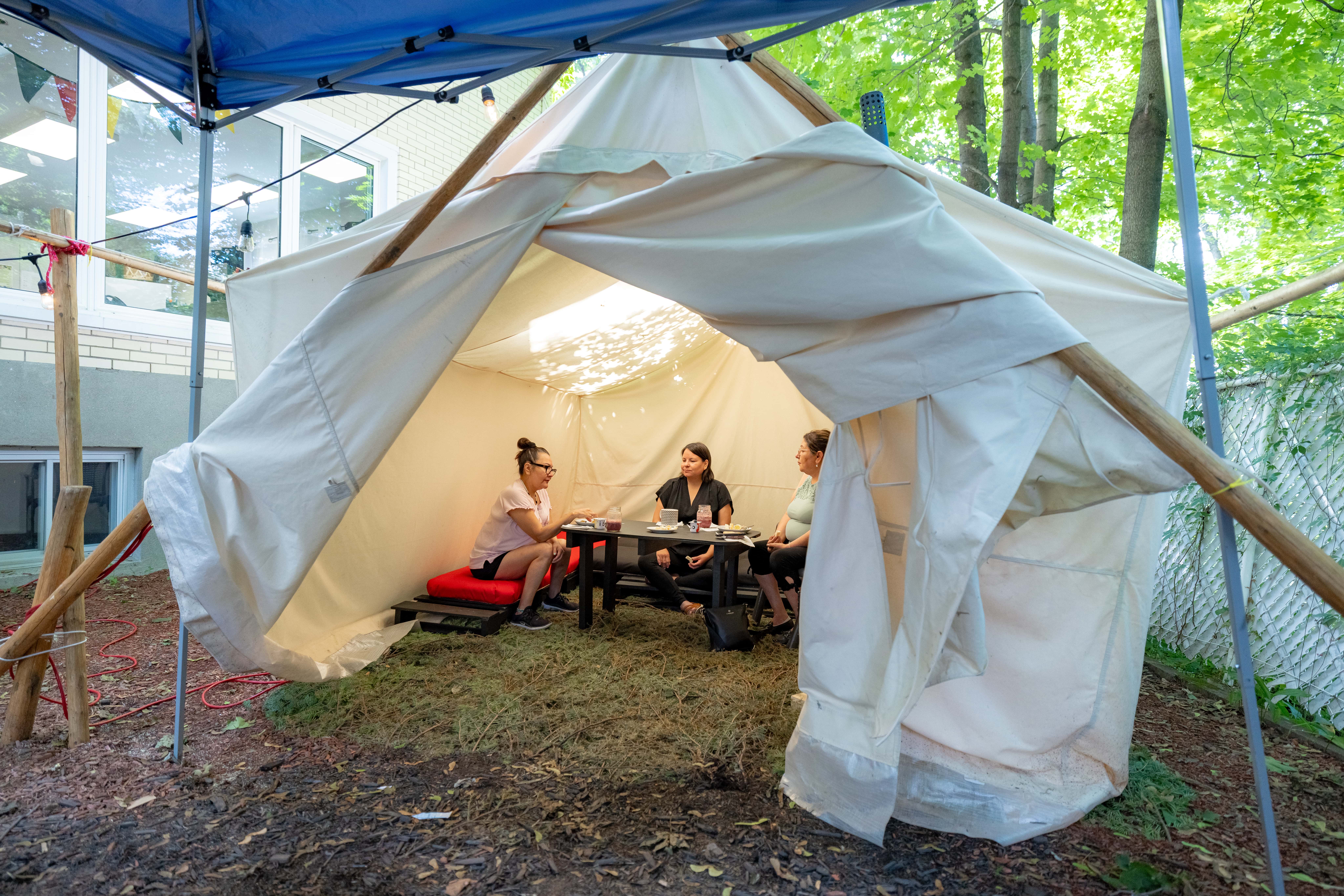Activité en plein air sous une tente dans la cour d’un Centre d’amitié autochtone, avec participants réunis autour d’une table.