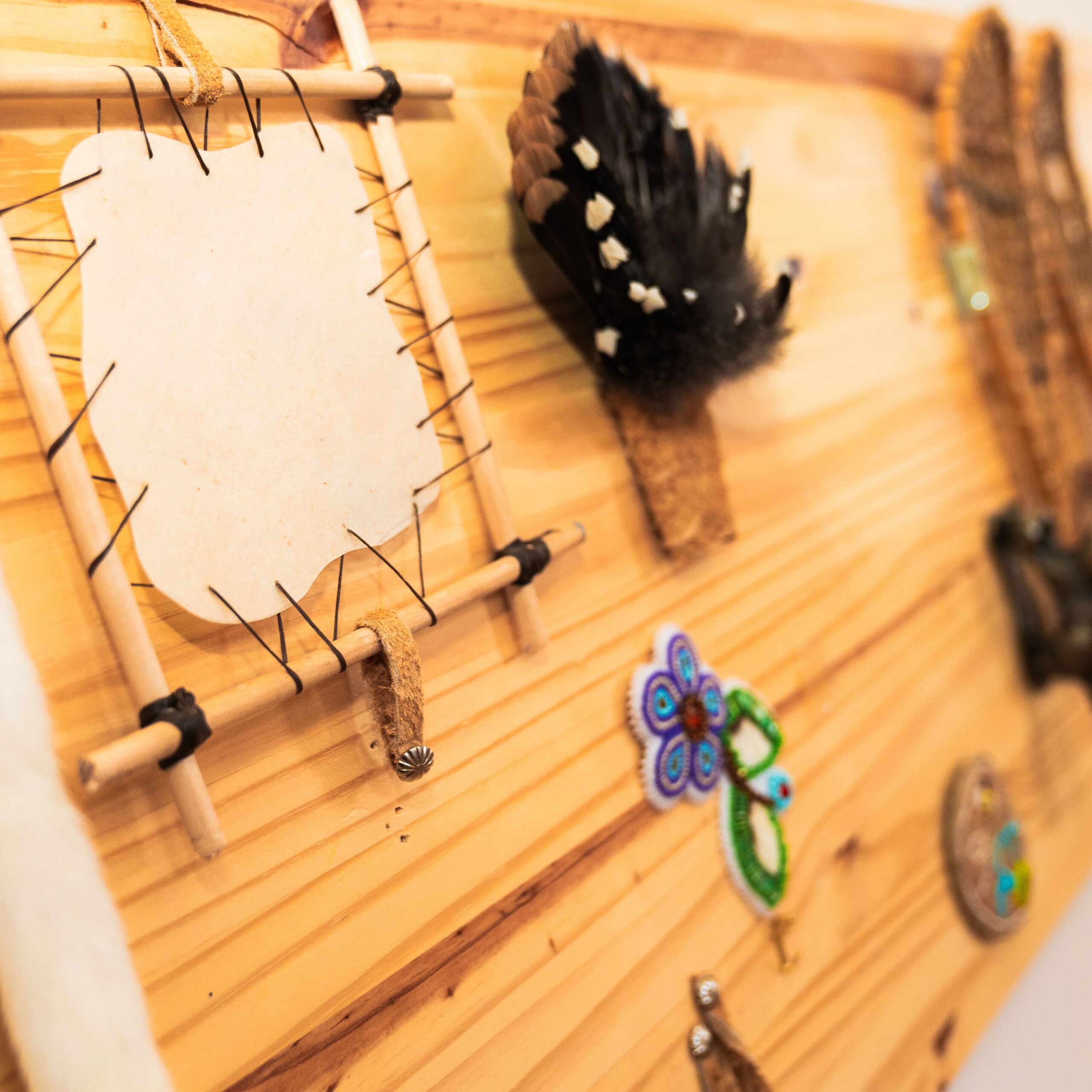 Planche en bois décorée d’éléments artisanaux autochtones : cadre en bois avec cuir, plumes et motifs perlés colorés.