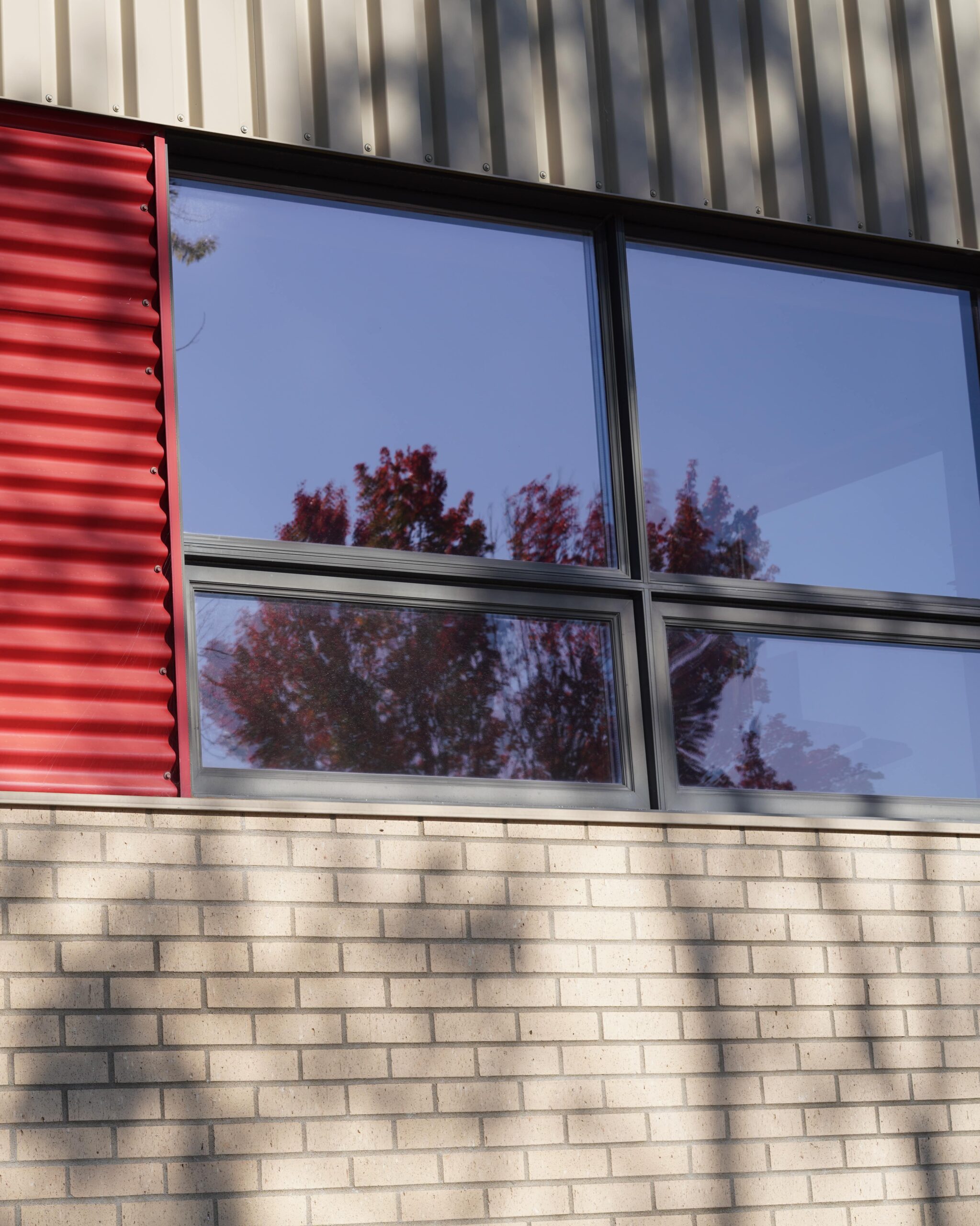 Façade d’un bâtiment en milieu urbain avec mur en briques pâles, panneau métallique rouge et grandes fenêtres reflétant des arbres.
