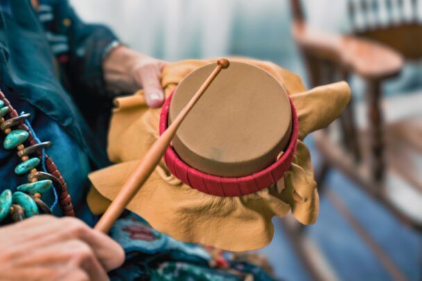 Main d'Ainée tenant un petit tambour autochtone recouvert de cuir avec bordure rouge et une baguette en bois, illustrant la pratique musicale traditionnelle.