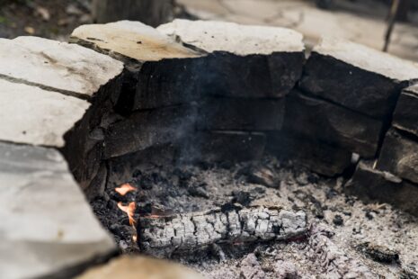 Foyer en pierres avec des braises et un morceau de bois partiellement consumé, illustrant un feu traditionnel en extérieur.