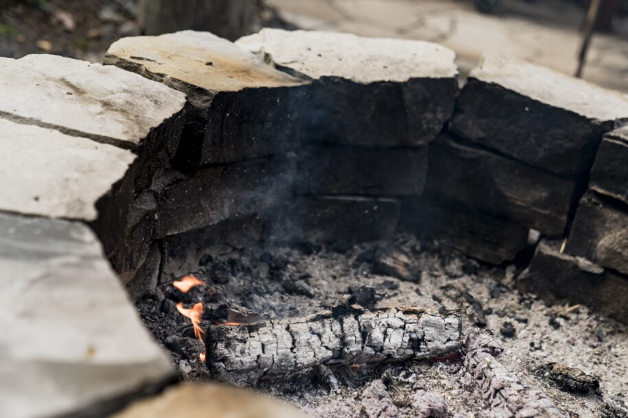 Foyer en pierres avec des braises et un morceau de bois partiellement consumé, illustrant un feu traditionnel en extérieur.