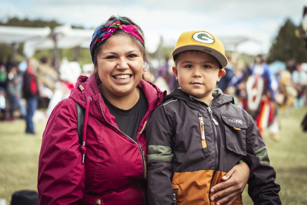 Mère autochtone tenant son enfant lors d’un pow wow en plein air, avec des participants et des cérémonies traditionnelles en arrière-plan.