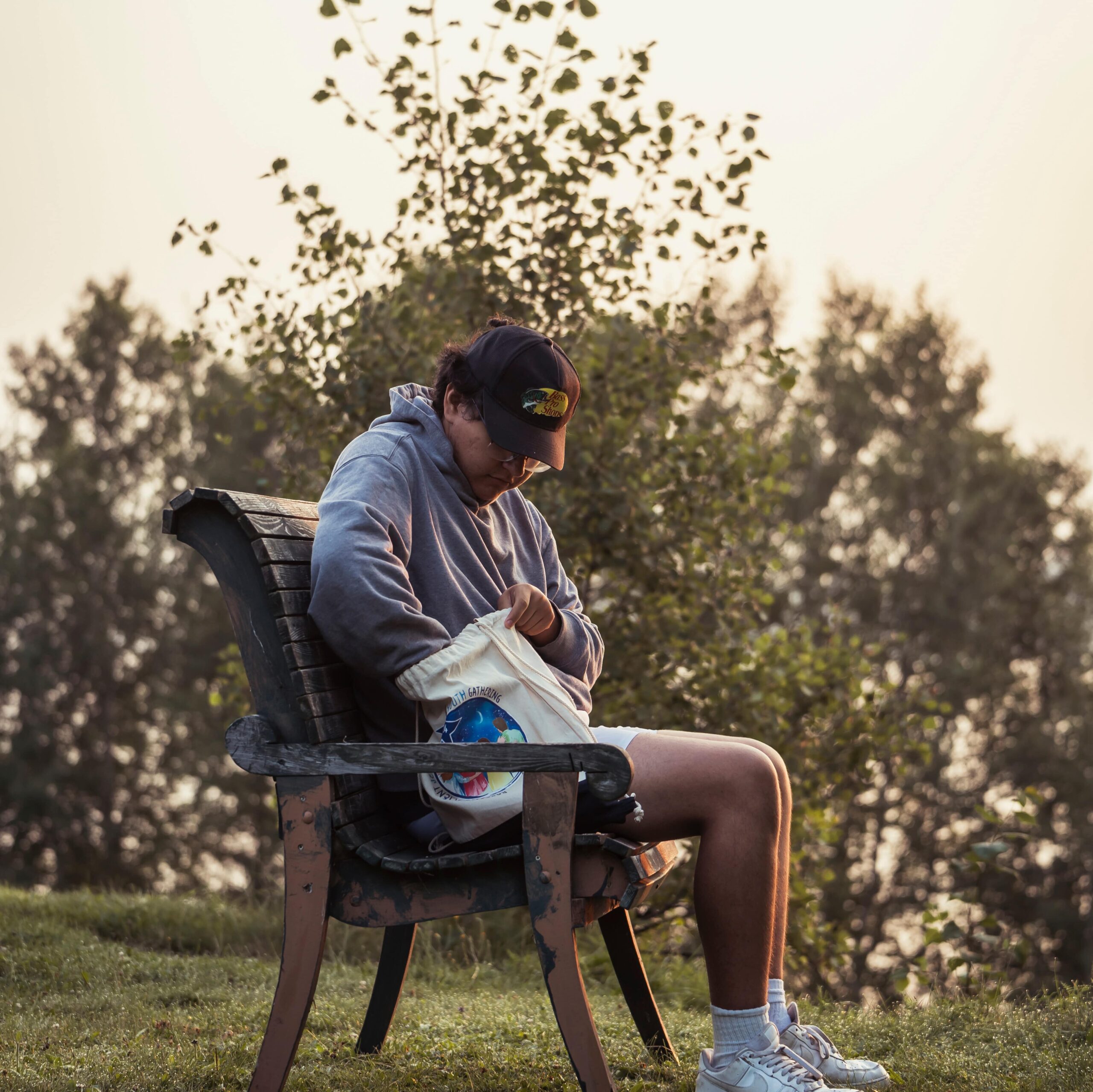 Personne autochtone assise sur un banc en bois à l’extérieur, fouillant dans un sac en tissu, avec des arbres en arrière-plan et une lumière douce de fin de journée.