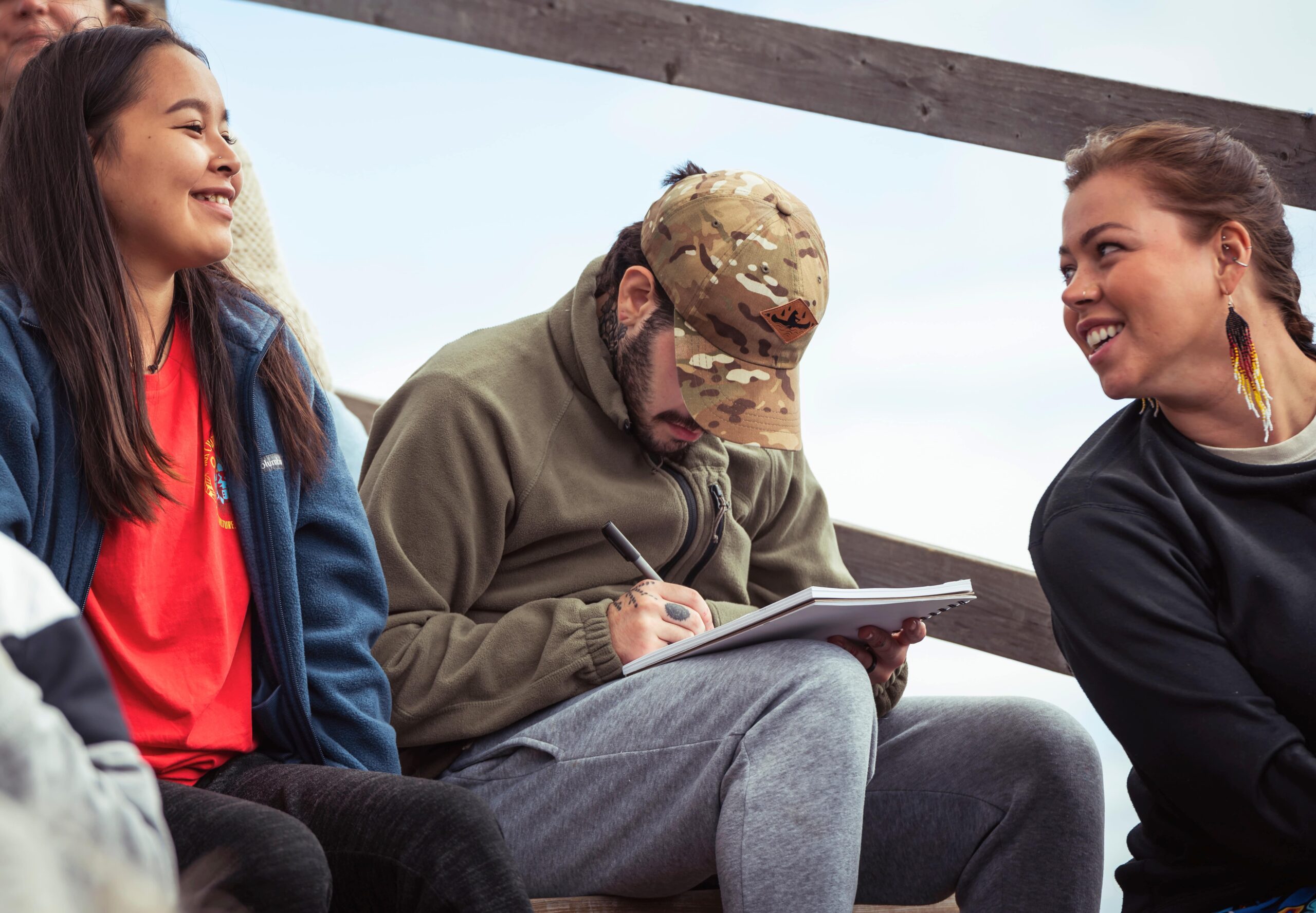 Personne assise sur des gradins en bois, écrivant dans un carnet avec un stylo, entourée d’autres personnes lors d’un événement en plein air