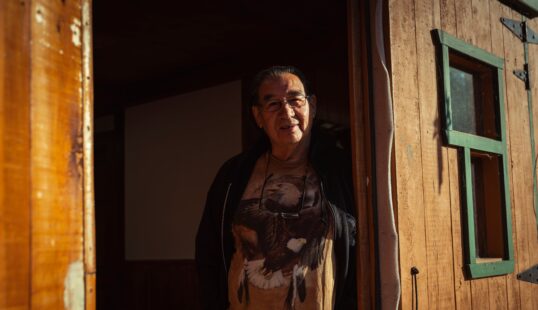 Aîné du mouvement des centres d’amitié autochtones se tenant dans l’embrasure d’une porte en bois, portant un chandail avec motif d’aigle –