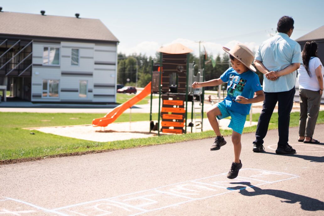 Enfant jouant à la marelle sur le terrain d’un milieu de vie de la Société immobilière du RCAAQ (SIRCAAQ) à Sept-Îles, avec modules de jeux et bâtiments résidentiels en arrière-plan.