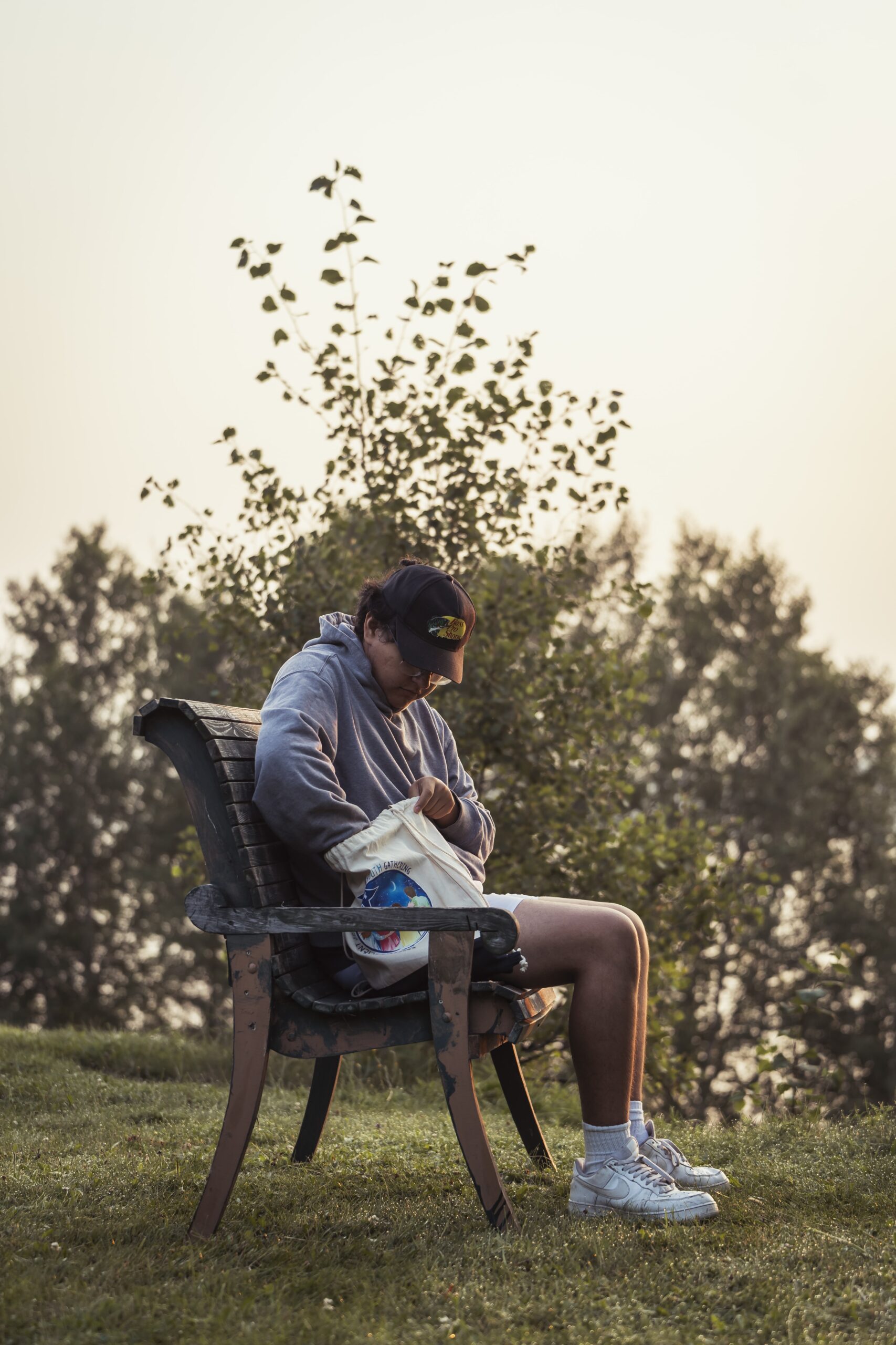 Personne autochtone assise sur un banc en bois à l’extérieur, fouillant dans un sac en tissu, avec des arbres en arrière-plan et une lumière douce de fin de journée.