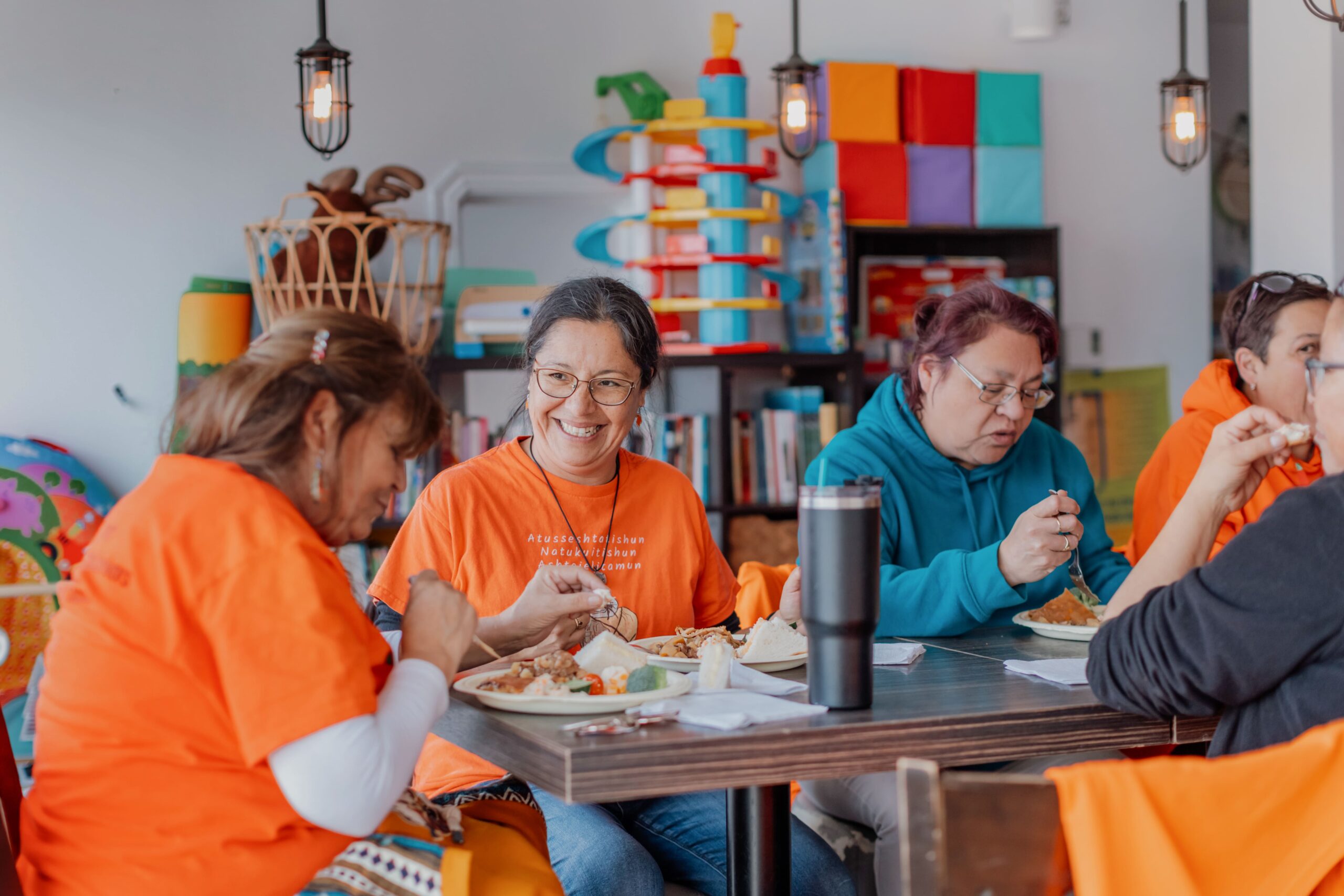 Repas communautaire partagé autour d’une table à l’intérieur d’un Centre d’amitié autochtone, avec assiettes de nourriture et décor coloré en arrière-plan.