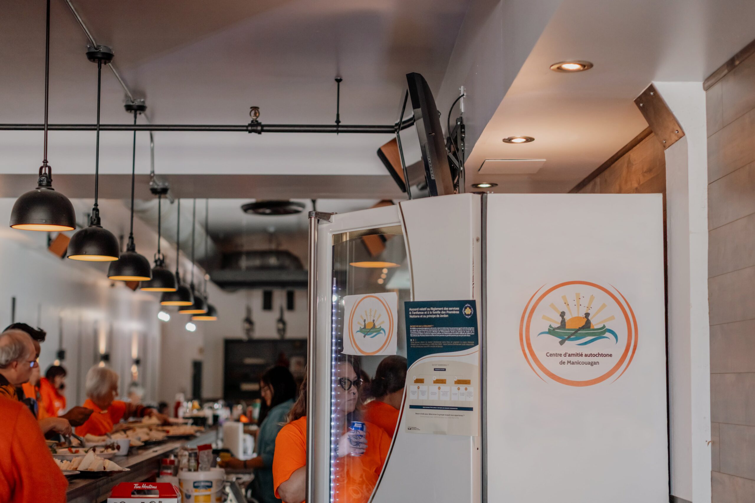 Intérieur du Centre d’amitié autochtone de Manicouagan avec comptoir de service, luminaires suspendus et logo du centre affiché sur un réfrigérateur.