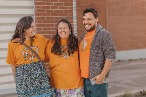 Trois membres d’un Centre d’amitié portant des chandails oranges, debout devant un mur de briques