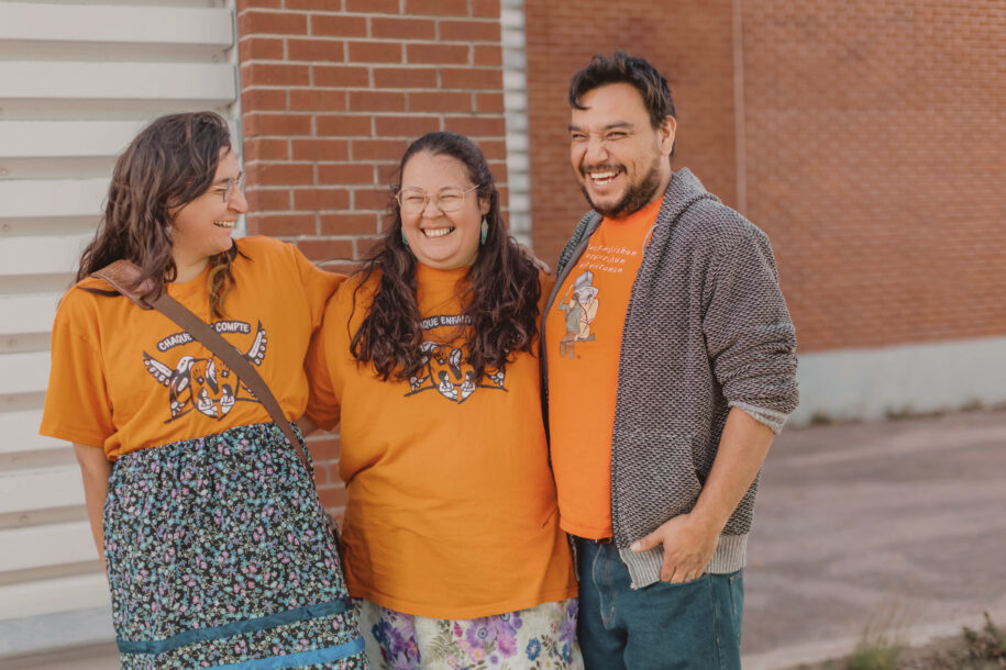 Trois membres d’un Centre d’amitié portant des chandails oranges, debout devant un mur de briques