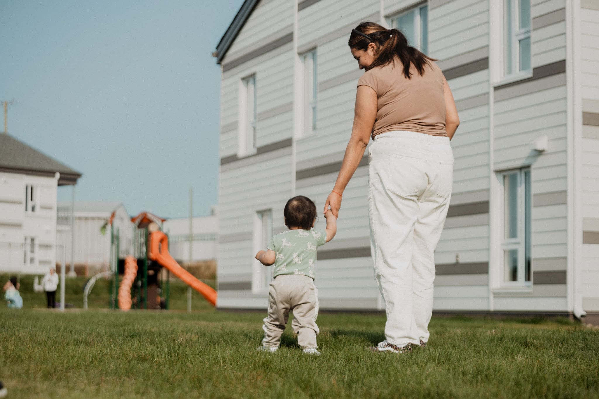 Personne marchant main dans la main avec un bébé sur une pelouse devant des bâtiments résidentiels et une aire de jeux dans un milieu de vie de la SIRCAAQ.